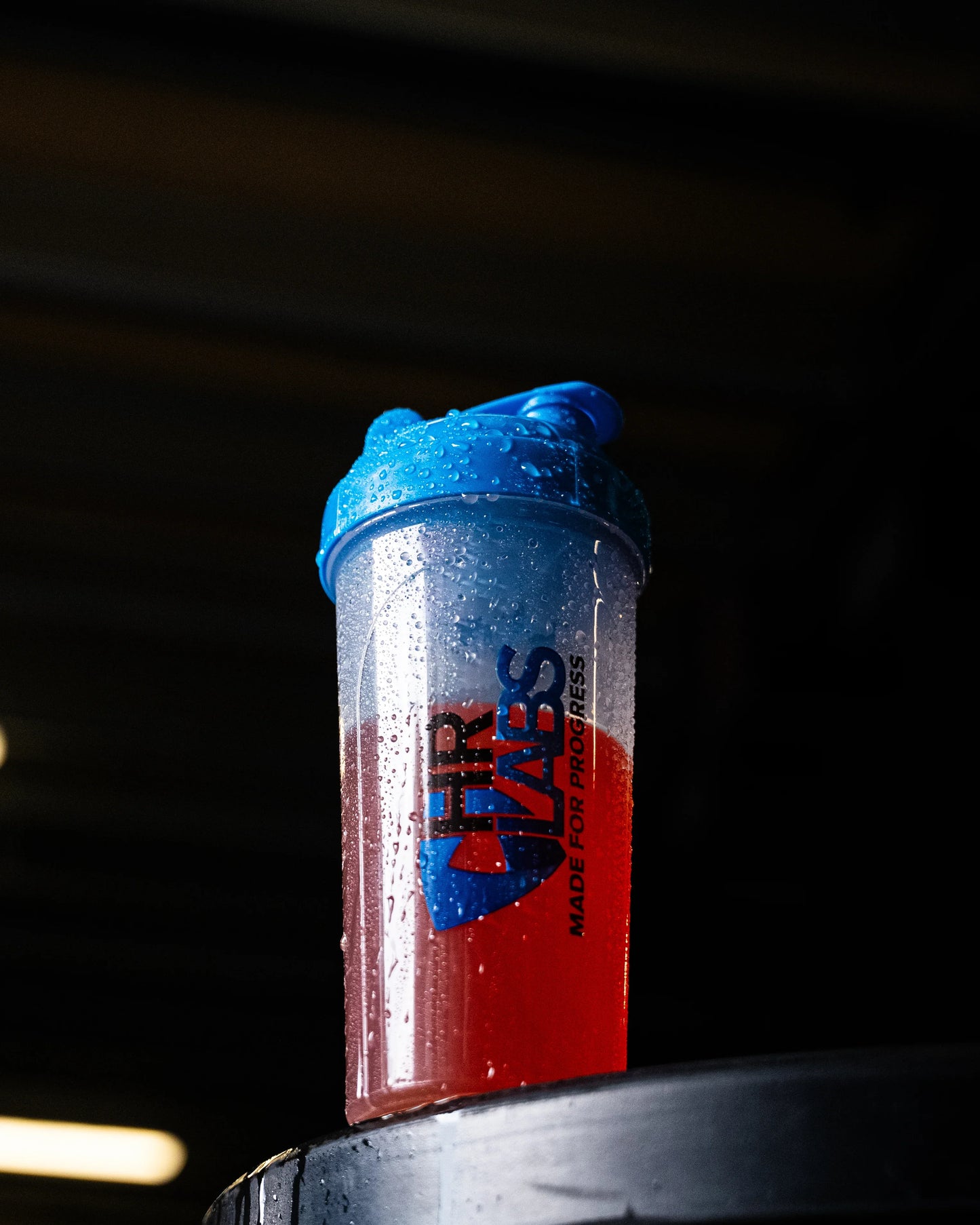 Shaker bottle with blue lid and red supplement drink, water droplets on surface